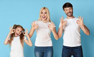 A smiling family of three, a young girl, a woman, and a man, stand side-by-side against a light blue background. All three are dressed in white shirts and jeans and are making the OK hand gesture photo