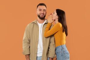 A woman in a yellow top leans in to whisper a secret to a smiling man wearing a beige jacket, all set against an orange backdrop. Both appear to be enjoying a playful and intimate moment. photo