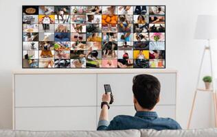 A man is sitting on a couch and watching television, holding the remote control in his hand. The TV screen is showing a grid of images, each depicting a different scene. photo