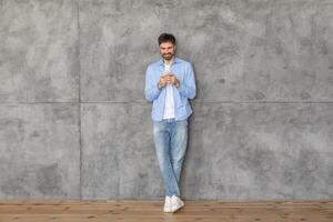 A young man in a blue shirt and jeans stands with his back against a concrete wall. He is using his smartphone and smiling as he looks down at the screen. The floor is made of wood. photo