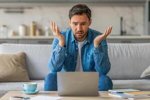 A man sits on a couch in his living room, looking confused as he stares at his laptop. His arms are raised in a gesture of frustration. A cup of coffee sits on a table in front of him. photo