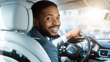 A smiling, African American businessman wearing a suit is driving a car. He is looking over his shoulder with a happy expression. The car is parked in a city and there is a blurred background photo