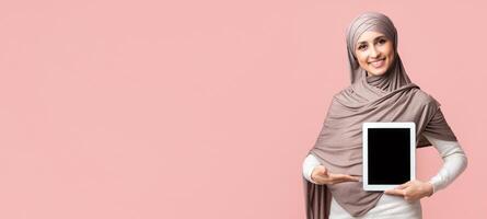 Young arabic woman in hijab holding and pointing at digital tablet with black screen, demonstrating new app or website over pink background photo