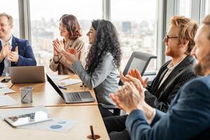 A group of business professionals sit around a table in a modern office, clapping their hands in appreciation. They are dressed in business attire and are engaged in a lively discussion. photo