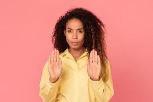 African American woman with curly hair is standing in front of a pink background, with both of her hands outstretched, palms forward, as if to say stop. photo