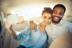 African American happy couple takes a selfie in the backseat of a car. The woman is holding a phone and smiling, while the man is smiling and has his arm around her photo