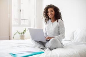 A Hispanic woman sits on a bed, focused on using a laptop computer. She types on the keyboard, with the screen displaying data. The room is well-lit, with a simple and clean decor. photo