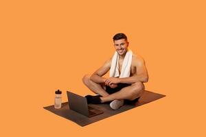 A fit man with a towel around his neck is seated cross-legged on a yoga mat, engaged in an online workout session on his laptop. A water bottle is placed next to him photo