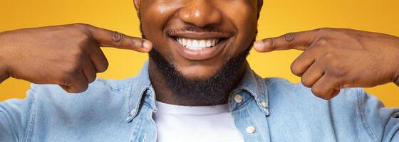 Dental health. Closeup of handsome black guy smiling wide and pointing at his perfect teeth, yellow background with copy space photo