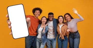 Five multiethnic friends stand together, smiling and posing enthusiastically, with one holding up a blank smartphone. The cheerful atmosphere is highlighted by a vibrant yellow backdrop. photo