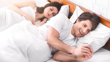 A man lies in bed next to his sleeping girlfriend, secretly looking at his phone while she sleeps. He is smiling mischievously, suggesting he is up to something. photo
