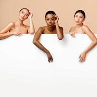 Three Multiracial Girls Posing Standing Naked Behind White Poster With Blank Space For Text On Gray Background. Studio Shot photo