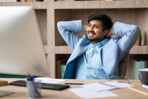 Indian young man in a light blue blazer and shirt is sitting at his desk with his hands behind his head. He is looking off to the side and smiling. There are documents and a keyboard on his desk photo