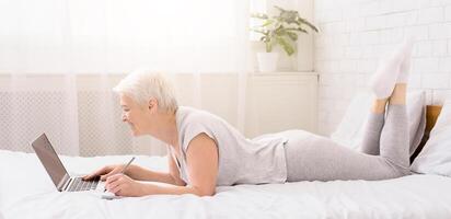 A senior woman is seen reclining on a bed, engaged with a laptop computer in front of her. She appears focused and intent on the screen, typing and scrolling, take notes photo