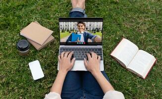 A student sits on the grass of a college campus, using a laptop to have a call with another student. The student on the laptop screen is smiling and holding a book. photo