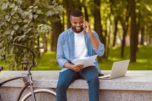 Handsome afro man sitting on bench and talking on cellphone while looking at documents in park, working out of office photo