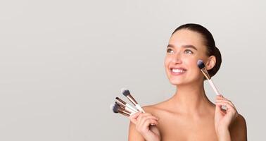 A woman smiling while holding a makeup brush in her hand. She appears happy and engaged in the act of applying makeup, empty space photo