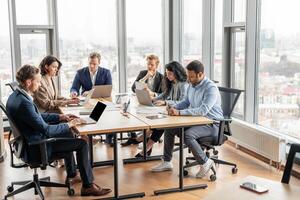 A group of business professionals work on their laptops at a large wooden table in a modern office. The office is filled with natural light from large windows overlooking a city. photo