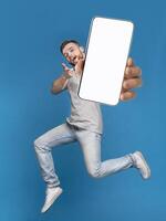 Emotional millennial redhaired guy pointing finger at camera, jumping with mobile phone on blue studio background, mockup for mobile app on white screen. Creative collage with website template photo