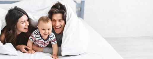 A happy family relaxes in a cozy bedroom, sharing delightful moments while playing under a fluffy blanket. The baby giggles joyfully, while parents smile at each other and their child, copy space photo