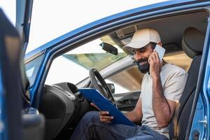 Indian man sits in the drivers seat of a blue car, engaged in conversation on his cell phone while holding a blue clipboard. He appears focused and engaged, with a relaxed demeanor photo