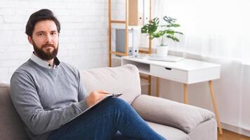 A man with a full beard is seated comfortably on a plush couch, holding clipboard. He appears relaxed and engrossed in his surroundings. photo