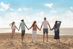 A group of five friends stands hand in hand on a sandy beach, enjoying a sunny day by the ocean waves with a clear blue sky above, back view photo