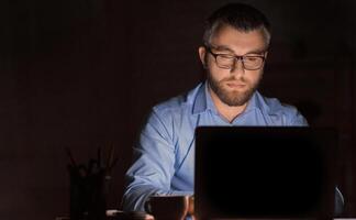 A man wearing glasses is intensely focused while working on his laptop in a dimly lit office environment during late-night hours. He appears to be deeply engaged in a task photo