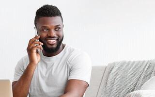 A young, African American man with a beard, wearing a white t-shirt, sits on a gray couch and smiles while talking on his phone. He is looking to the side as he speaks on the phone photo