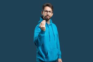 A man in a vibrant blue hoodie stands against a solid dark background, extending his hand in a gesture that suggests he is inviting or requesting something from the viewer. photo
