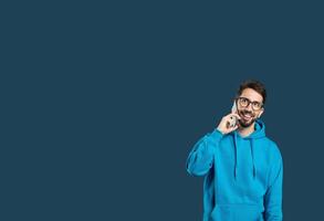 A man wearing a blue hoodie stands against a dark background, smiling while talking on his smartphone. He appears cheerful and engaged in conversation, showcasing a relaxed demeanor, copy space photo