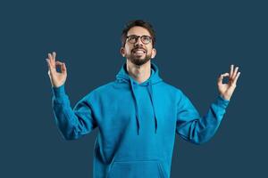 A man is practicing mindfulness meditation, wearing a blue hoodie and glasses while maintaining a serene expression. The background is a calming shade of blue, enhancing the peaceful atmosphere. photo