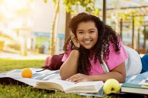 A cheerful student lies on a blanket in a park, engrossed in a book. Surrounded by fruits and greenery, she enjoys a relaxing afternoon under the sun. photo