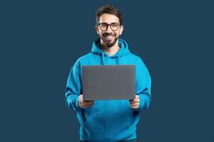 A young man wearing a bright blue hoodie smiles widely while holding a laptop in front of him. The backdrop is a simple dark color that highlights his joyful expression and tech device. photo