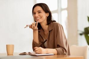 Happy female manager talking on speaker phone using cellphone, writing memo in notebook, looking aside, copyspace photo