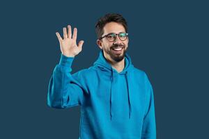 A cheerful young man greets with a smile and raised hand, dressed in a blue hoodie, set against a dark, solid backdrop. His happy expression conveys friendliness and warmth. photo