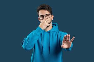 A young man in a blue hoodie stands against a solid dark background, pinching his nose with one hand while extending the other hand in a stopping gesture, indicating a strong smell. photo
