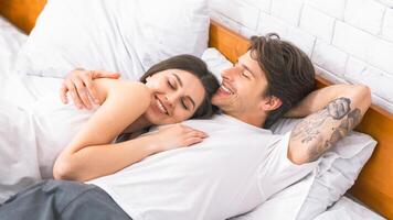A young couple is lying in bed together, both smiling and seemingly enjoying their time together. The woman is nestled into the mans side, her head resting on his chest, and he has his arm around her. photo