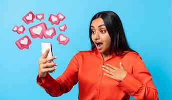 Amazed Indian millennial woman texting lover on smartphone, expressing wow emotion on blue background. Modern digital communication, dating mobile app concept photo