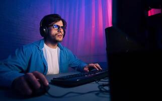 A young man sits in front of a computer, wearing headphones and blue light glasses. He is typing on the keyboard with his right hand and using a mouse with his left hand. photo