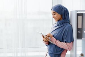 Business Application. Smiling Muslim Secretary Using Digital Tablet While Standing Near Window In Modern Office, Copy Space photo