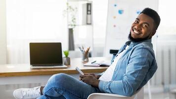 Portrait of cheerful african american employee using smartphone at workplace, browsing internet or messaging with friends and looking at camera photo