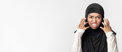 Hate Concept. Angry arabic woman in hijab emotionally grimacing and gesturing in rage and looking at camera, posing over light background, closeup photo