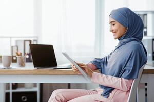 Technologies For Business. Smiling Islamic Businesswoman Using Digital Tablet While Sitting At Workplace In Modern Office, Side View With Free Space photo