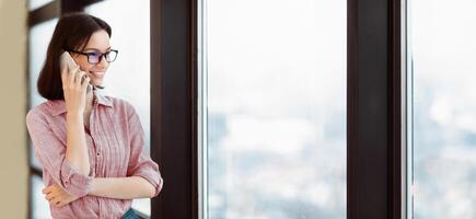Communication Concept. Portrait of female student in specs talking on cellphone, looking out the window, copyspace photo
