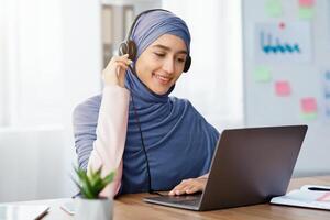 Customer Representative Service Concept. Portrait Of Smiling Female Call Center Operator In Hijab And Headset Using Laptop In Office photo