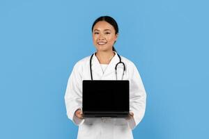 A smiling Asian woman healthcare worker in a white lab coat holds a laptop against a blue backdrop, symbolizing the integration of technology in patient care and medical practice. photo