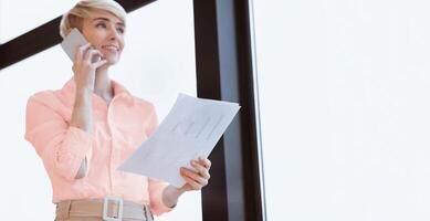 Business Call. Businesswoman Talking On Cellphone Holding Folder Standing Near Window In Modern Office. Selective Focus, Low Angle photo