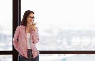 Young female manager in specs talking on speaker phone using cellphone, standing by window in office, copyspace photo