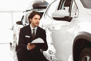 Writing Report. Serious handsome male assistant sitting near car and filling insurance form photo